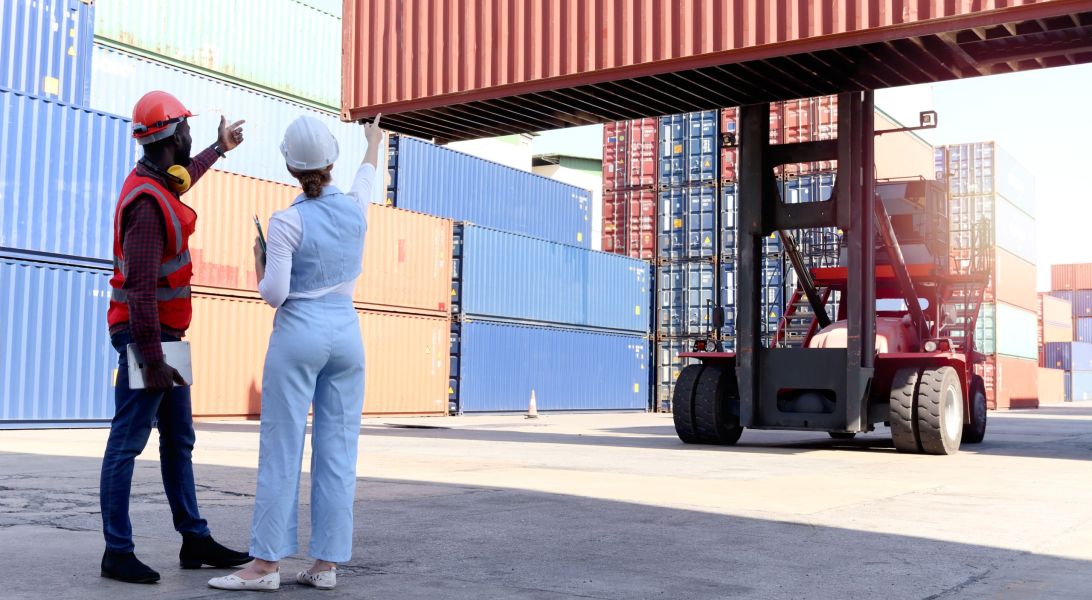 Behind two workers wear safety vest and helmet discussing at log Behind two workers wear safety vest and helmet discussing at logistic shipping cargo container yard. African American engineer man talk with beautiful young woman boss with blonde hair at workplace.
